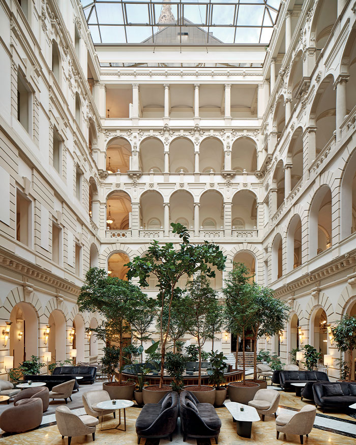 A glass-roofed atrium lobby looks over four levels of
Neo-Renaissance arcades, with evening light warming the pilasters while cafe seating gathers around a central grove and marble inlay floor; Photographs courtesy Anantara New York Palace Budapest Hotel