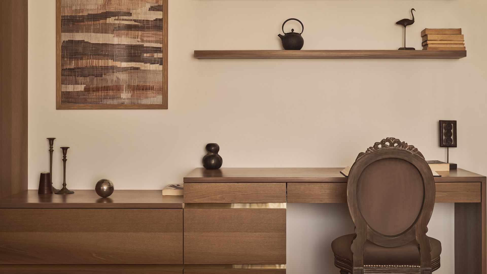 At the study corner in the master bedroom, a wooden table takes centere stage, its top polished to a soft, golden gleam that catches the light, while the front grain is rustic and textured under the fingertips, full of character. The chair, upholstered in a deep, comforting brown, invites you to sink into it. On the table, tiny trinkets rest, and above, two wooden slabs double as shelves, holding books, clocks, and keepsakes, their raw warmth echoing the room’s calm. The beige wall behind hosts a framed artwork, adding just the right pause for the eyes; Photography by Yadnyesh Joshi