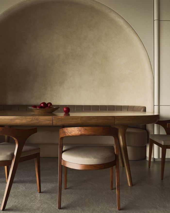 A gentle plaster arch curves overhead, softening the space and framing the dining area. The palette glows in honeyed beiges, warm woods, and soft browns, while the table, chairs, and banquette seem to melt into one another; Photography by Yadnyesh Joshi