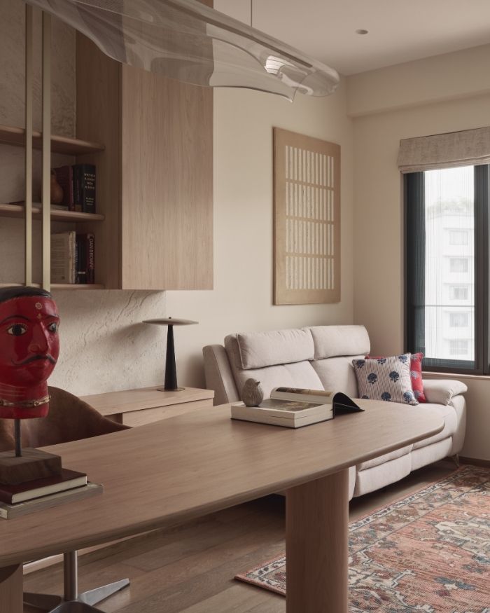 Sunlight pours into the study, giving the space a calm, welcoming feel. By the window sits a small, cream-colored sofa, perfect for curling up with a book, with a piece of artwork hanging just above. Next to it, a simple console table holds a lamp, and a wooden shelf above keeps books neatly within reach. Ahead, a wooden desk is set with a few books and another piece of artwork, while a delicate, transparent hanging sculpture floats above. The walls are a soft beige, the carpet adds warmth underfoot, and the stone-finish wall behind the desk quietly takes centere stage. Wood and veneer from ZZ Decor, along with curtains and upholstery from Bharat Furnishings, tie the whole room together into a space that feels light, warm, and lived-in; Photography by Yadnyesh Joshi