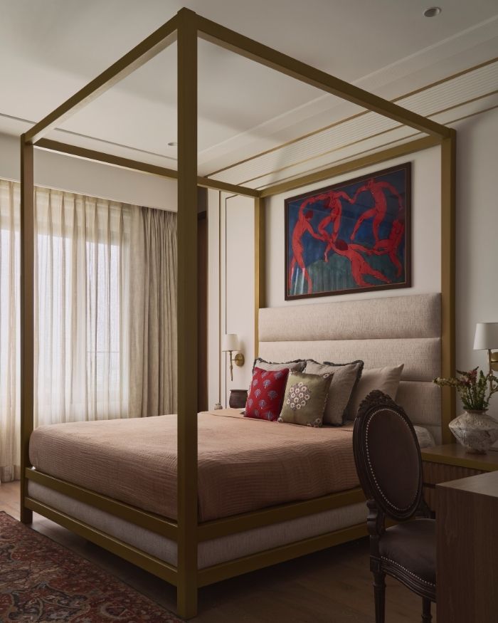 Soft light filters through delicate curtains, spilling gently across the room and resting on the four-poster bed upholstered in a tender pink by Sarita Handa and cushions from Cottons & Satins. Above the headboard, a striking painting in red and blue draws the eye, adding drama to the serene white walls. At the foot of the bed, a warm, intricately woven rug by Obeetee invites bare feet to linger, while the engineered wood flooring adds natural warmth underfoot; Photography by Yadnyesh Joshi