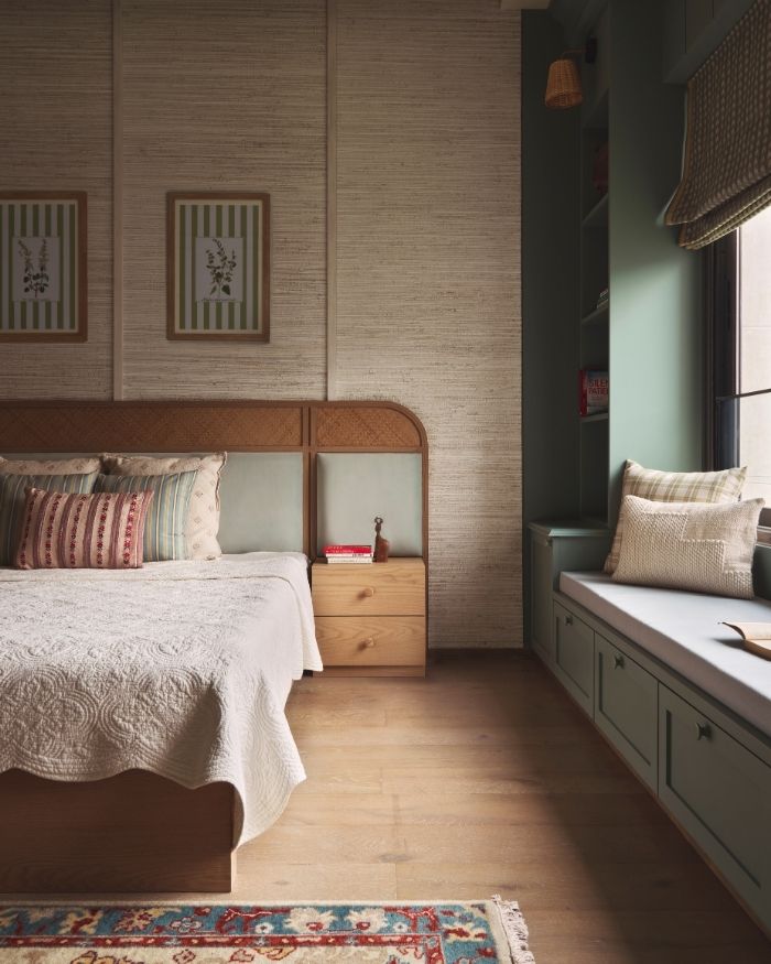 The window nook beckons with soft sage-green upholstery, a quiet spot where you could curl up with a book or watch the light shift across the room. Next to it, a matching cabinet holds daily objects out of sight, its colour blending gently with the nook. The bed sits softly in the frame, its cane headboard topped with a painting that adds a dash of colour. At the foot, the carpet feels warm underfoot, its texture inviting you to pause and sink in; Photography by Yadnyesh Joshi