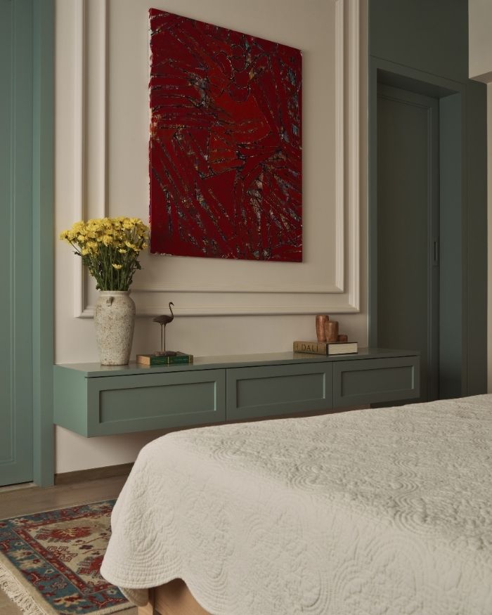 This corner of the guest bedroom unfolds in shades of sage green, white and a striking pop of red. The soft white upholstery of the bed by Sarita Handa peeks into view, but the eye is drawn to the wall by the foot of the bed: a bold red painting anchors the space, while sage-green drawers beneath hold a small collection of books, a ceramic vase brimming with sunny yellow flowers, and a few carefully chosen decorative accents. To the right, a matching sage wardrobe adds a calm, grounded presence; Photography by Yadnyesh Joshi