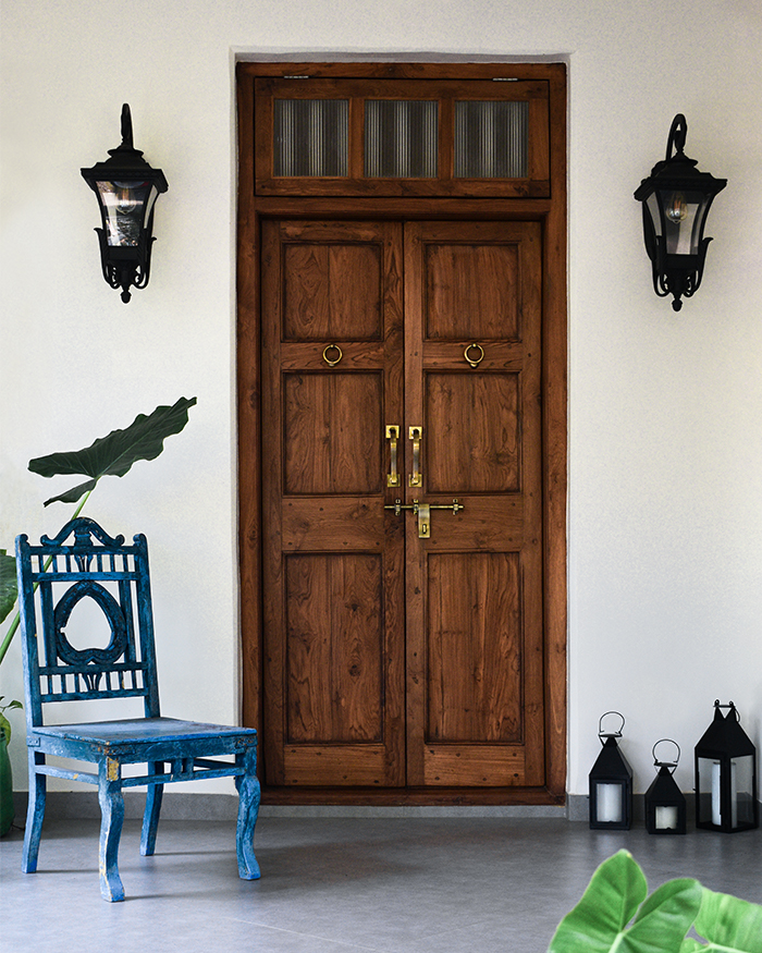 A teakwood door with brass fittings sets the tone, framed by lanterns from a local vendor. A 107-year-old blue chair and lush greenery add layers of memory; Photography by Studio Marés