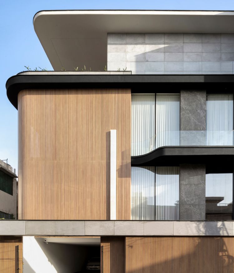 The street facade pairs vertical timber battens by Natural Veneers with grey stone cladding by Nexion; a dark steel trim separates the two surfaces. The cantilevered slab sweeps outward in a slow curve, its underside painted black; Photography by Avesh Gaur