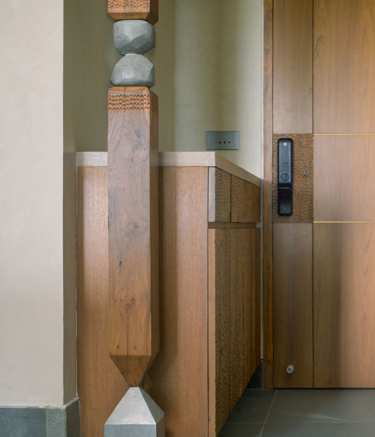 The signature entry column by Grava Stones - Kota stone and solid teak - with hand-carved wood banding, embossed detailing and a faceted geometric stone base; the natural wood and hammered-surface entry door unit is visible behind. Photography by Hemant Patil
