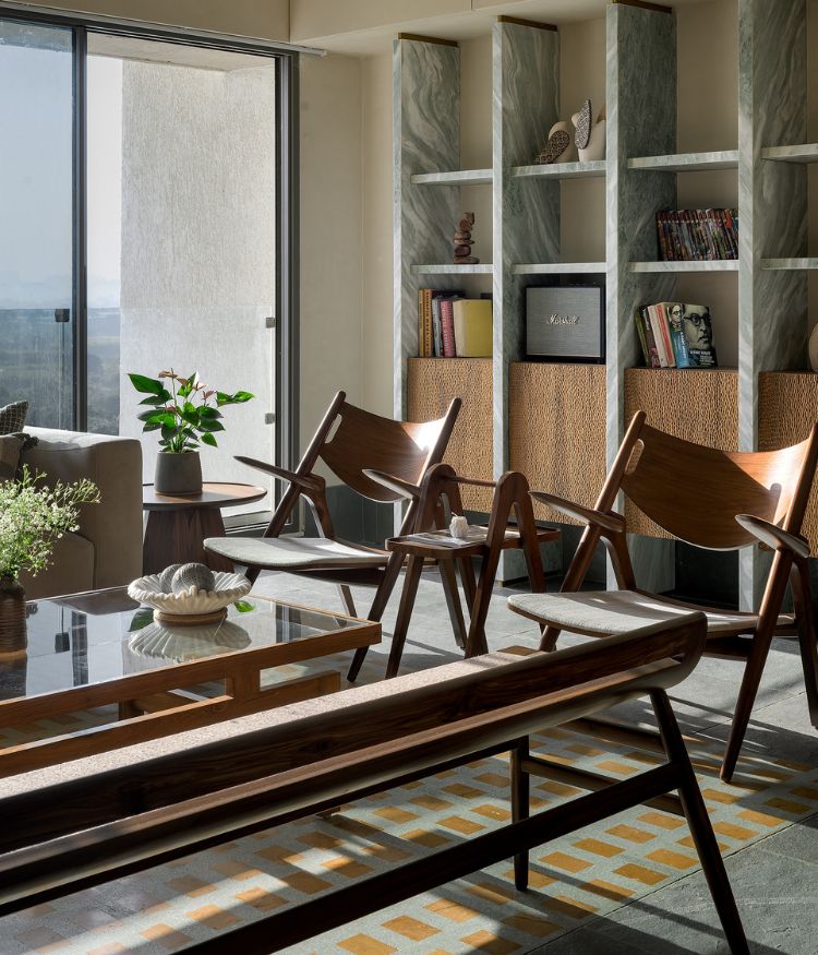 Looking towards the custom bookshelf unit - verde green marble-framed with woven cabinet fronts - across the living room; Designbee teak armchairs sit on the Jaisalmer terrazzo panel by Stonecasa; the city unfolds through the full-height window to the left. Photography by Hemant Patil