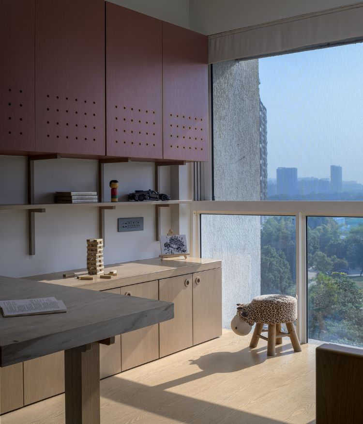 The child's room: birch plywood cabinetry and deep maroon perforated upper units line the walls; a concrete-top study desk runs below; a giraffe stool sits by the full-height window overlooking the city skyline. Photography by Hemant Patil