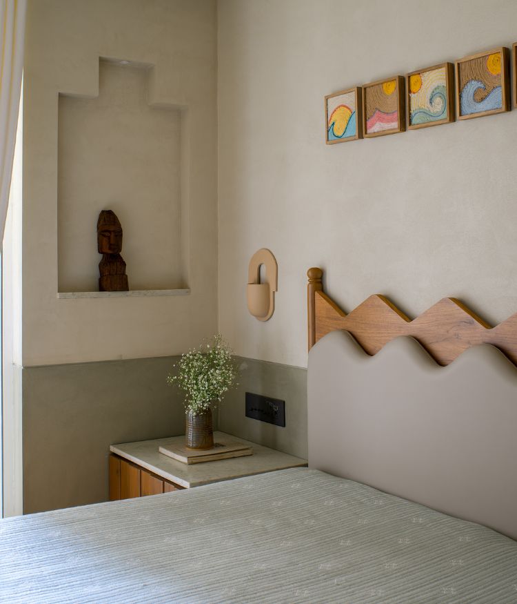 The bedroom's built-in plaster niche houses a carved wood sculpture; a marble-top teak side table holds a woven vase with baby's breath; wave-motif artwork panels are arranged above the zigzag oak headboard by Spaceace Design Co; wall sconce by Harshita Jhamtani Designs. Photography by Hemant Patil
