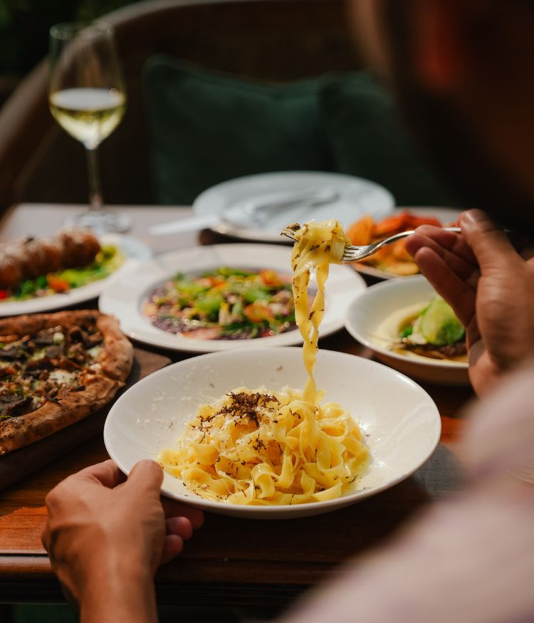 Bowl of creamy pasta being twirled with a fork at a table set with pizza, wine, and Italian dishes