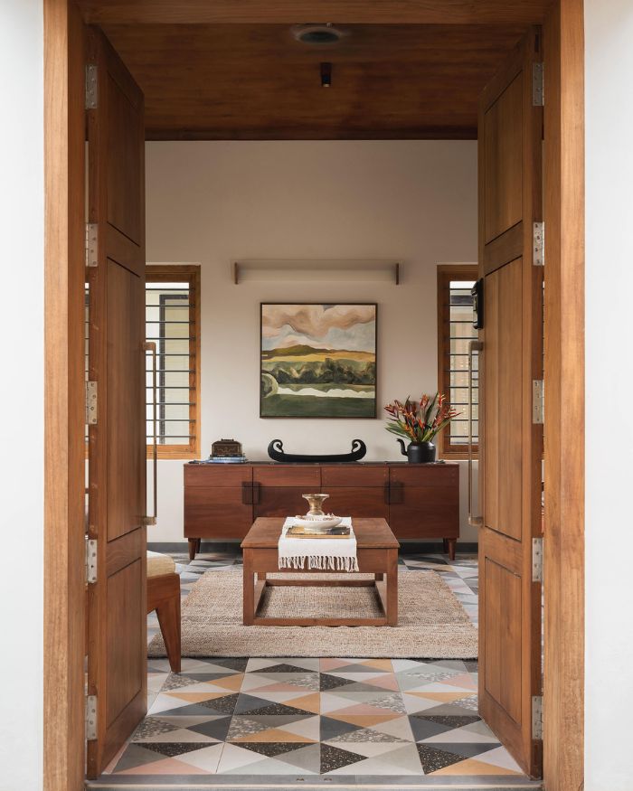 The entry is framed by a generous teak door surround — a wooden sideboard by local craftsmen holding artefacts from Fort Kochi, a landscape artwork above it, and a low coffee table on a jute rug setting the room's unhurried register. Styled by Fymin Naif and Nimitha Harith; Photography by Justin Sebastian and Athul Niranjan