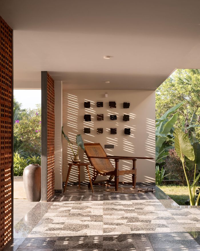 A planter's chair by local craftsmen rests in the afternoon shadow of the terracotta jaali screen, striped light falling across the granite floor. An artefact from Fort Kochi sits beside it; a grid of dark ceramic wall pieces by local craftsmen holds the wall behind. Styled by Fymin Naif and Nimitha Harith; Photography by Justin Sebastian and Athul Niranjan