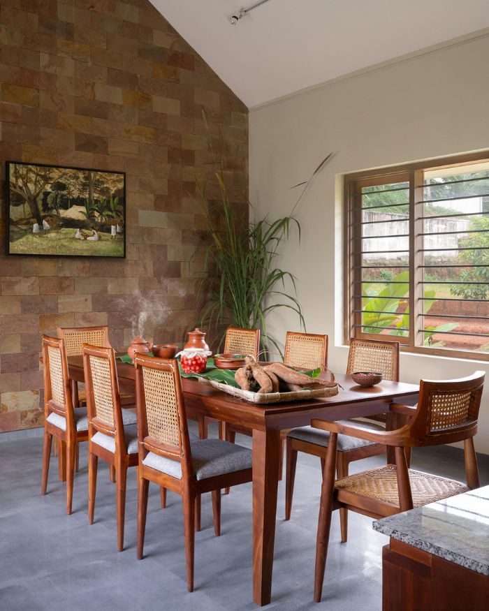 The dining room rises into a pitched ceiling, a laterite stone wall anchoring the space with earthy warmth. A landscape painting from Espravo presides over the table by local craftsmen, cane-back chairs lining either side, a garden window letting Kerala green into the far corner. Styled by Fymin Naif and Nimitha Harith; Photography by Justin Sebastian and Athul Niranjan