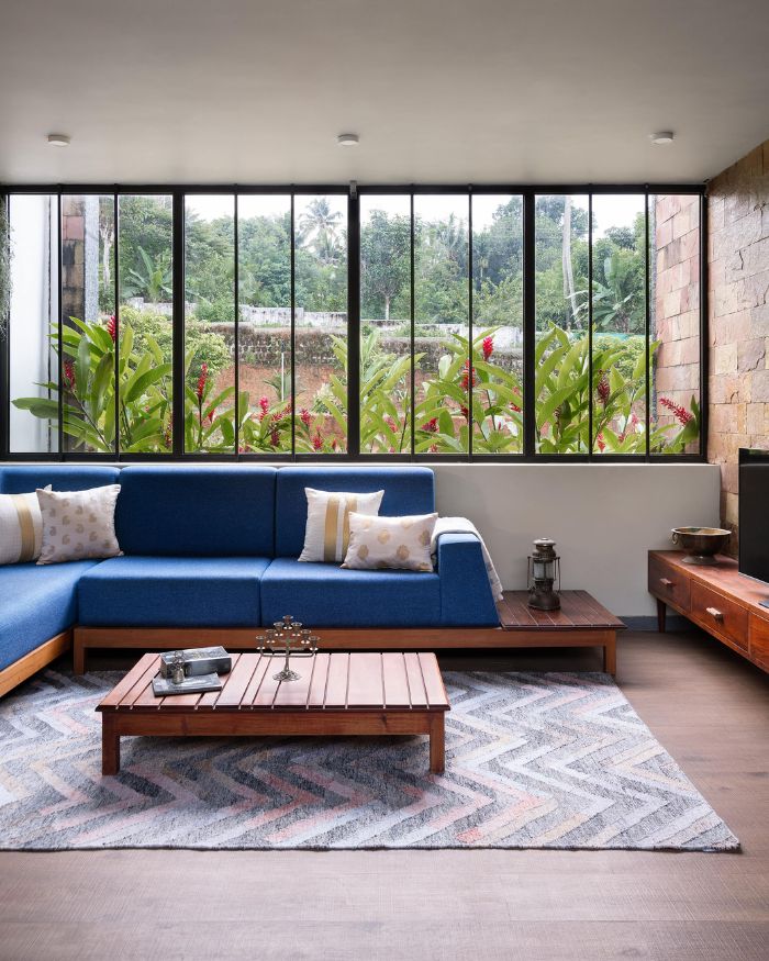 A cobalt blue sofa by local craftsmen faces the garden through a full-width picture window, red heliconia blooms pressing close against the glass. A slatted teak coffee table and chevron rug anchor the seating, a laterite stone accent wall framing the television to the right. Styled by Fymin Naif and Nimitha Harith; Photography by Justin Sebastian and Athul Niranjan