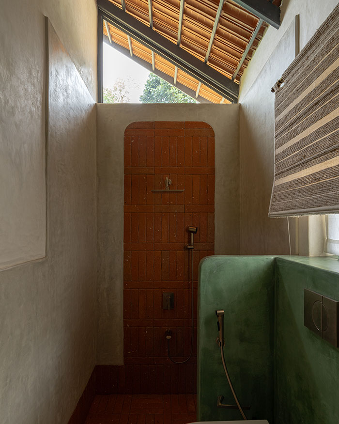 Lit from above, the bathroom pairs exposed brick with smooth plaster surfaces and a curved green partition; Photography by Syam Sreesylam
