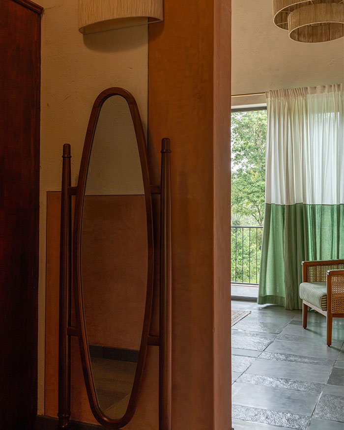 A transition space opens toward the balcony. An oval mirror and glimpses of greenery extend the interiors outward; Photography by Syam Sreesylam