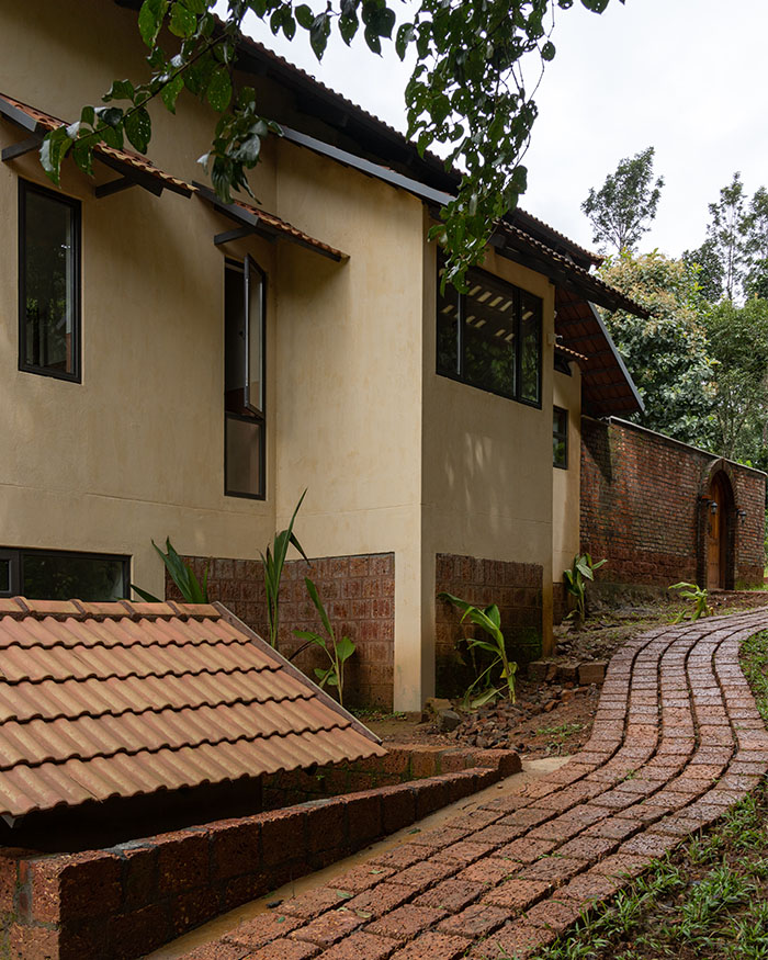 A brick path curves along the edge of the home, leading past walls and shaded openings toward a tucked-away entrance; Photography by Syam Sreesylam
