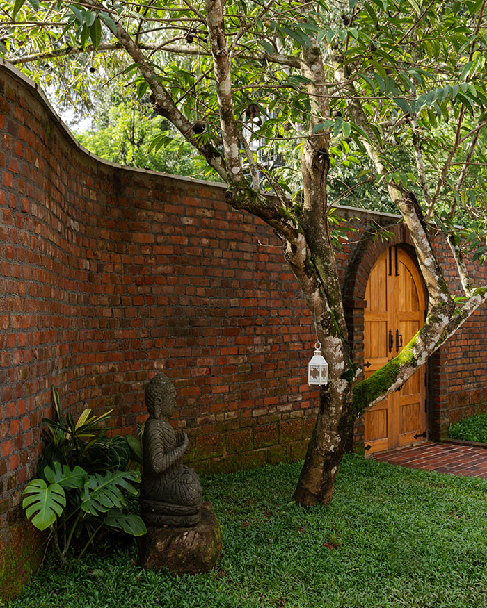 Within the curved brick enclosure, a courtyard gathers around a tree. The carved stone figure and arched doorway add layers of texture; Photography by Syam Sreesylam