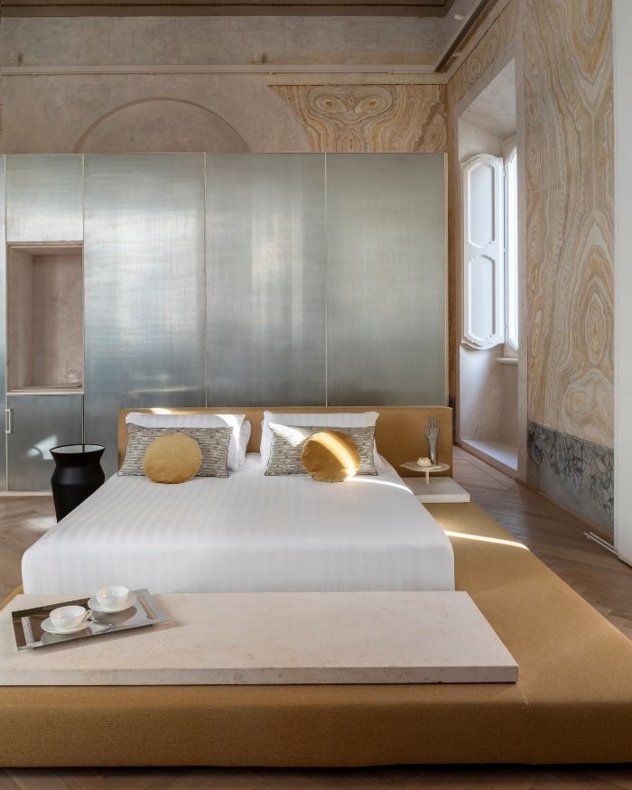 A rich interplay of veined stone, brushed metal and warm timber defines the bedroom, where daylight filters through period windows to accentuate the space’s sculptural material palette. The composition is anchored by a custom Letto bed based on a design by architect Boragina, framed by expressive stone surfaces and a monolithic metallic backdrop that lends the room a quietly dramatic presence; Photography by Serena Eller of Eller Studio