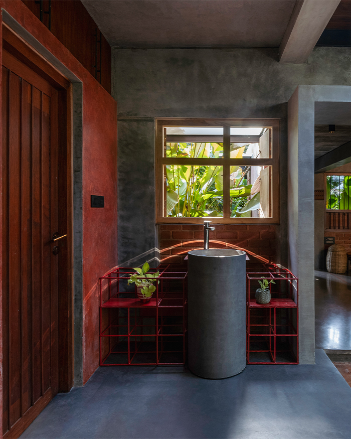 A monolithic pedestal basin stands against a backdrop of exposed concrete, brick and custom red steel shelving, reflecting the home’s raw material palette; Photography by Ar. Prashanth Mohan of Running Studios