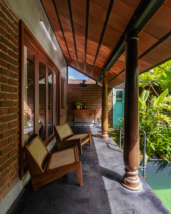 The shaded verandah, framed by timber columns and exposed brick walls, opens gently onto the lush tropical landscape beyond; Photography by Ar. Prashanth Mohan of Running Studios