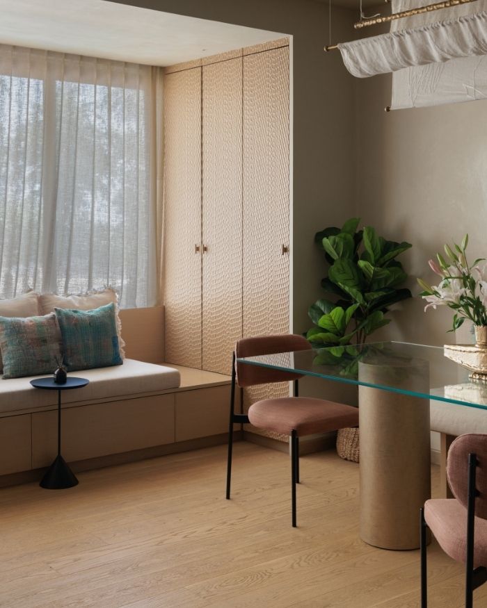 Textured wardrobe shutters and a cushioned window bench frame the dining zone, where kantha cushions and a fiddle-leaf fig soften the warm oak and neutral plaster; Photography by Wabi Sabi Studio