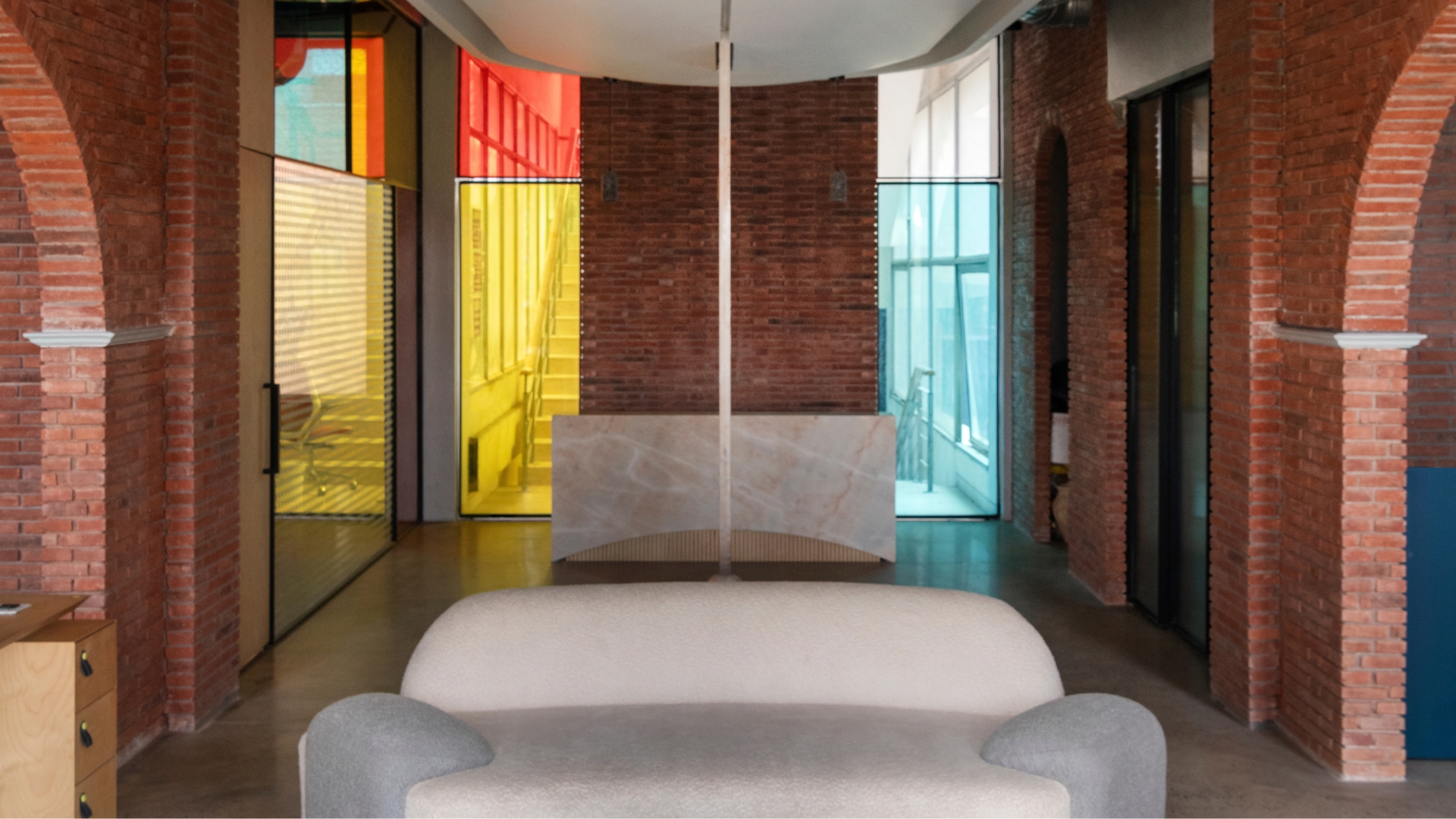 A grey bouclé sofa and stone-topped coffee table sit beneath a sweeping plaster ceiling, framed by glimpses of the red, yellow and blue glazed cabins beyond